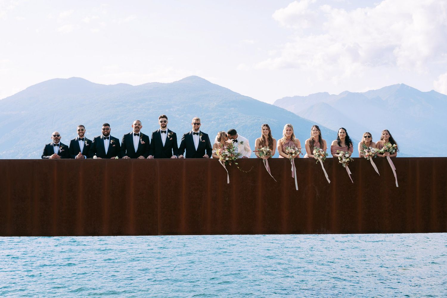 A group of people sit together on a wooden ledge overlooking a scenic mountain lake view.