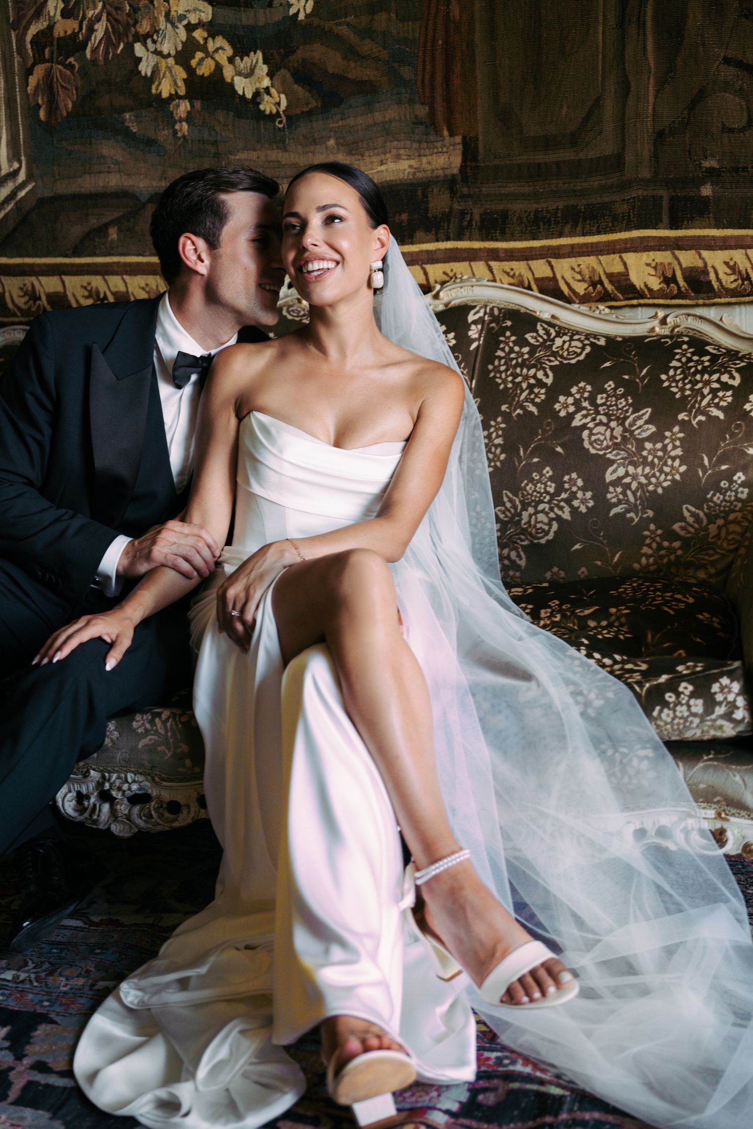 A romantic wedding couple lounges on an ornate vintage sofa with flowing white veil and pink dress details.