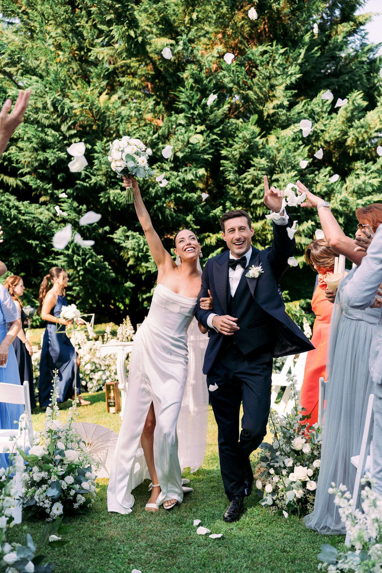 Wedding party celebrating outdoors by throwing white flower petals in joyful celebration during summer ceremony.