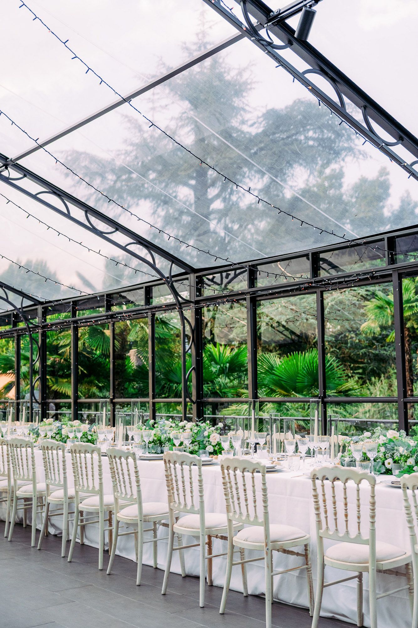 Clear tent wedding reception venue with elegant white chairs arranged in rows under a transparent roof with lush greenery outside.