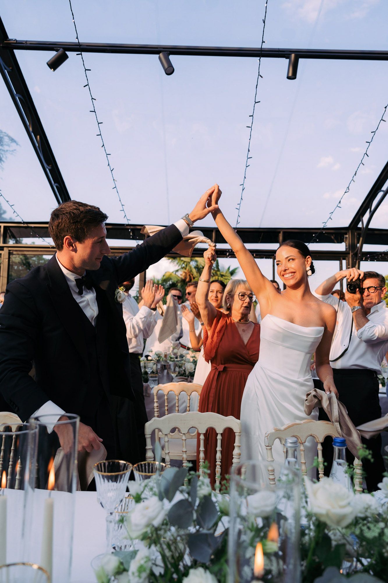 Wedding guests dance and celebrate under a clear tent decorated with string lights and floral arrangements.