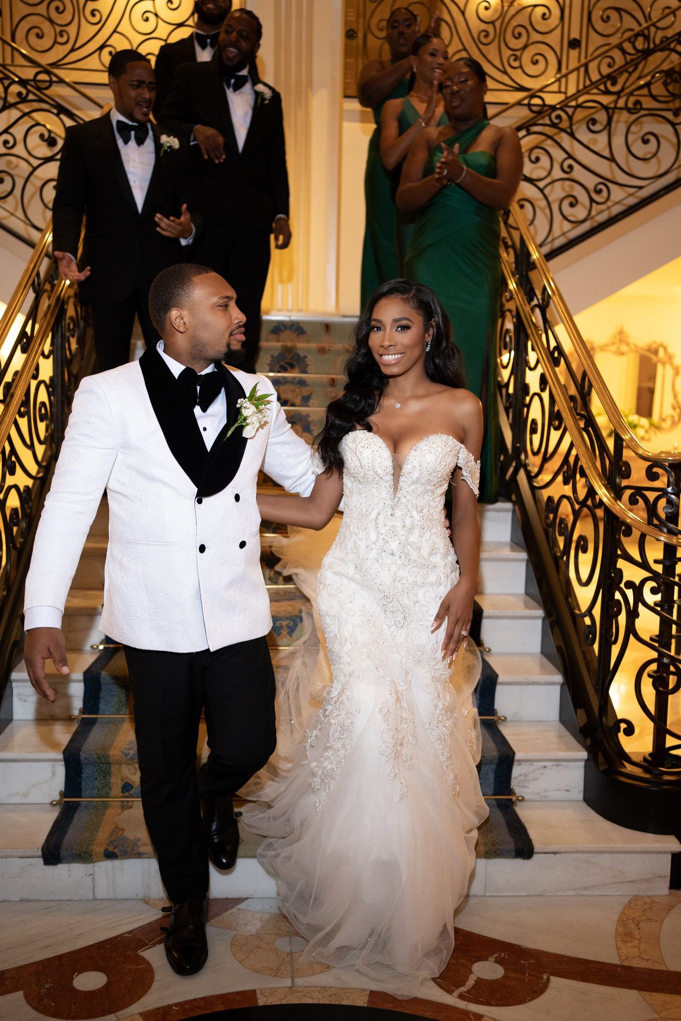 A couple descends an elegant staircase at a formal event, with the groom in white tuxedo and bride in lace mermaid gown.