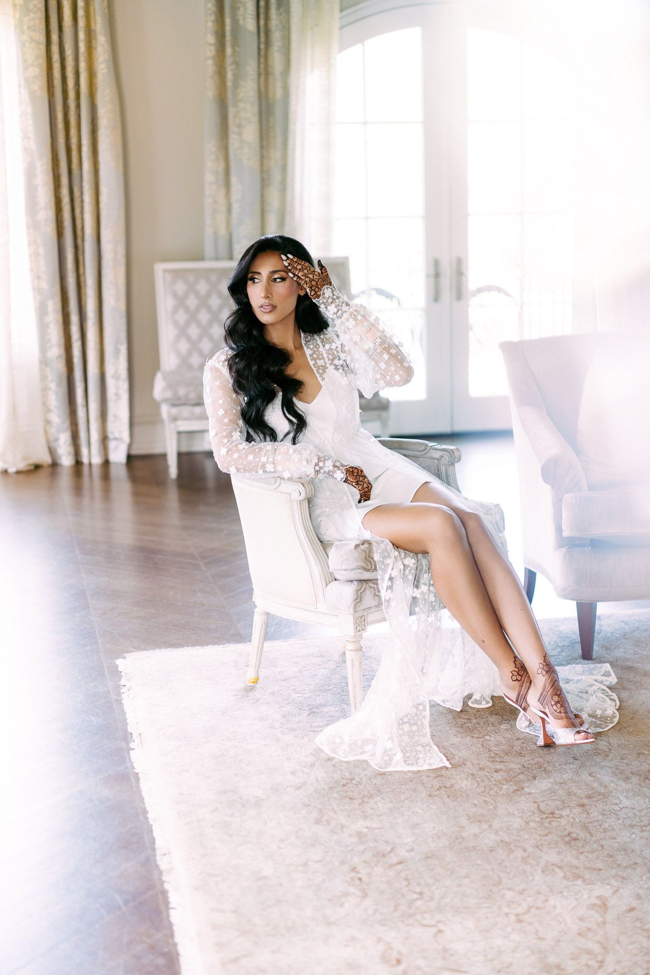 A bride in a white lace robe sits elegantly on a luxurious chair in a bright, airy room with flowing curtains.