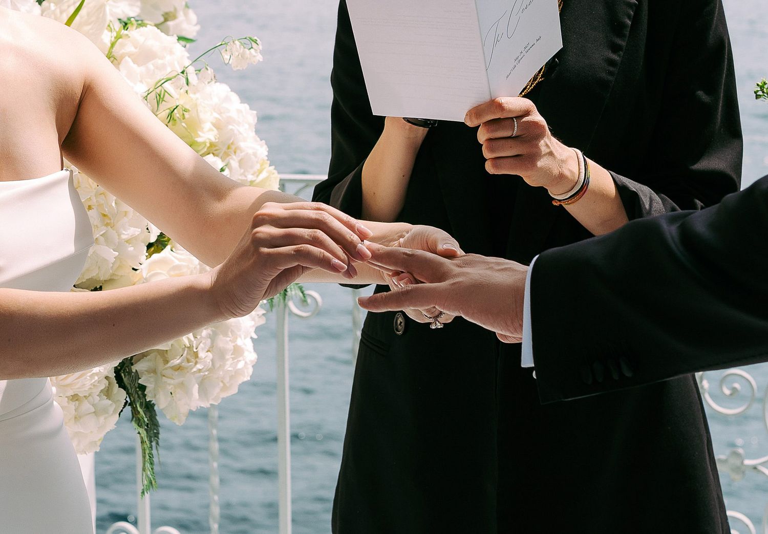 Close-up of hands exchanging wedding rings during outdoor waterfront ceremony with white floral bouquet.
