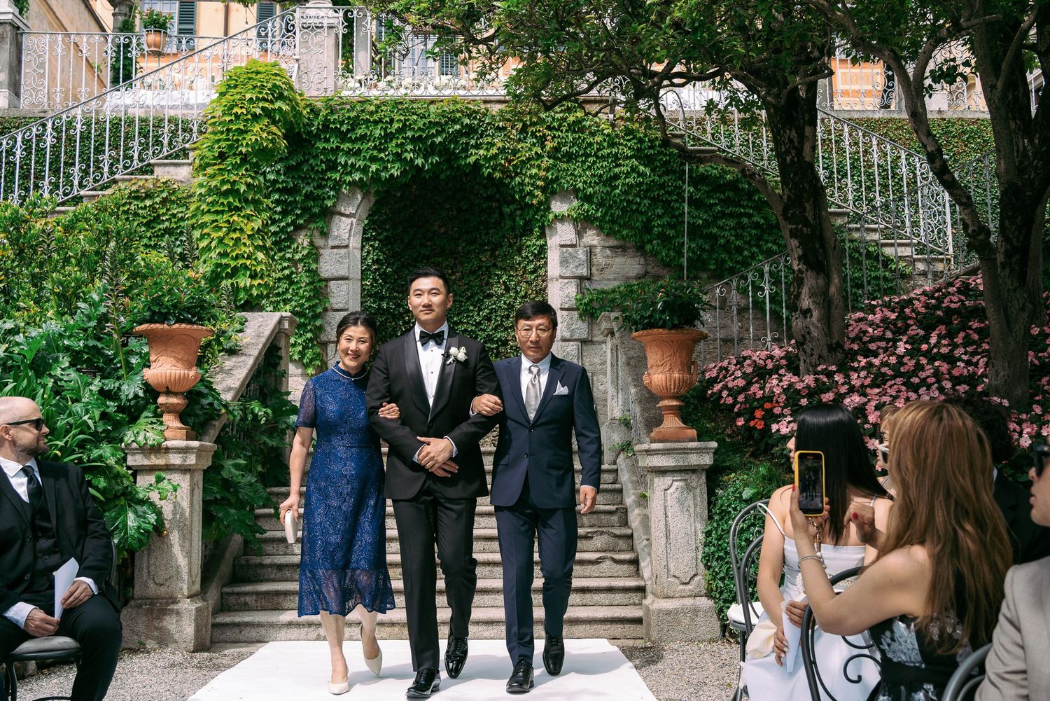 Wedding guests gather in a lush garden setting with stone architecture and greenery creating an elegant outdoor atmosphere.