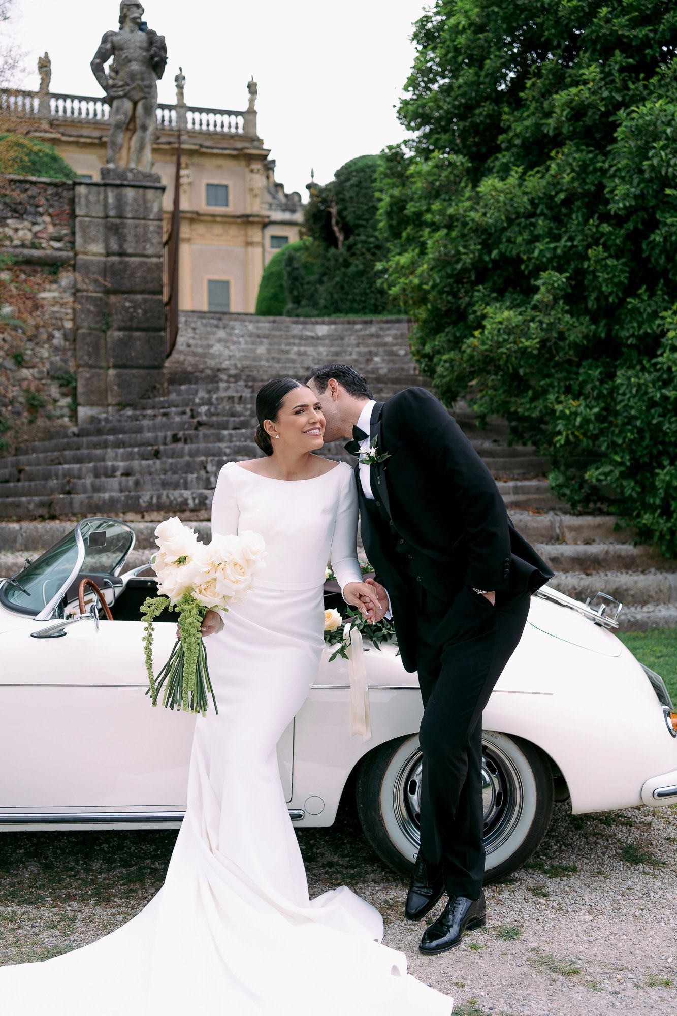 A couple poses by a vintage white car during their wedding photoshoot at a historic estate.