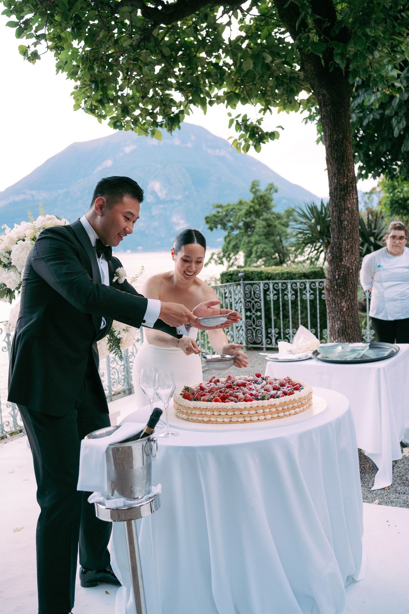 Wedding guests serve themselves dessert from elegant white-clothed tables with a scenic mountain and lake backdrop.