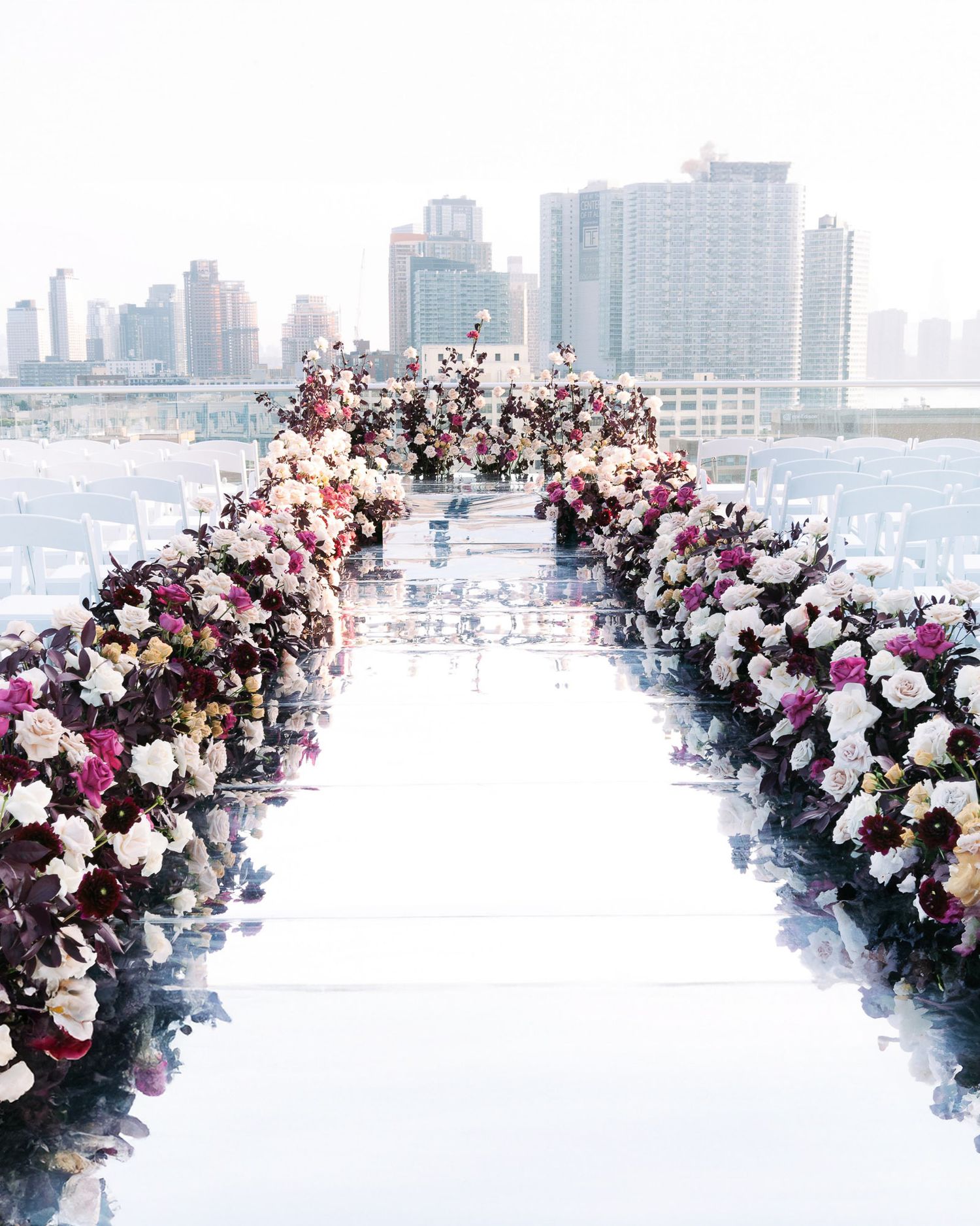 A panoramic floral arch installation with pink and white blooms frames a city skyline view across a body of water.