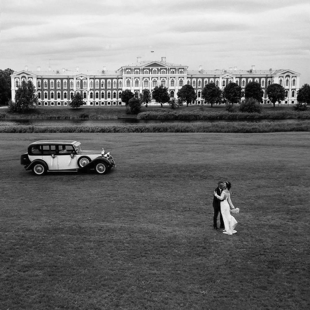 A vintage car drives across a vast green lawn in front of a grand historical building in black and white.