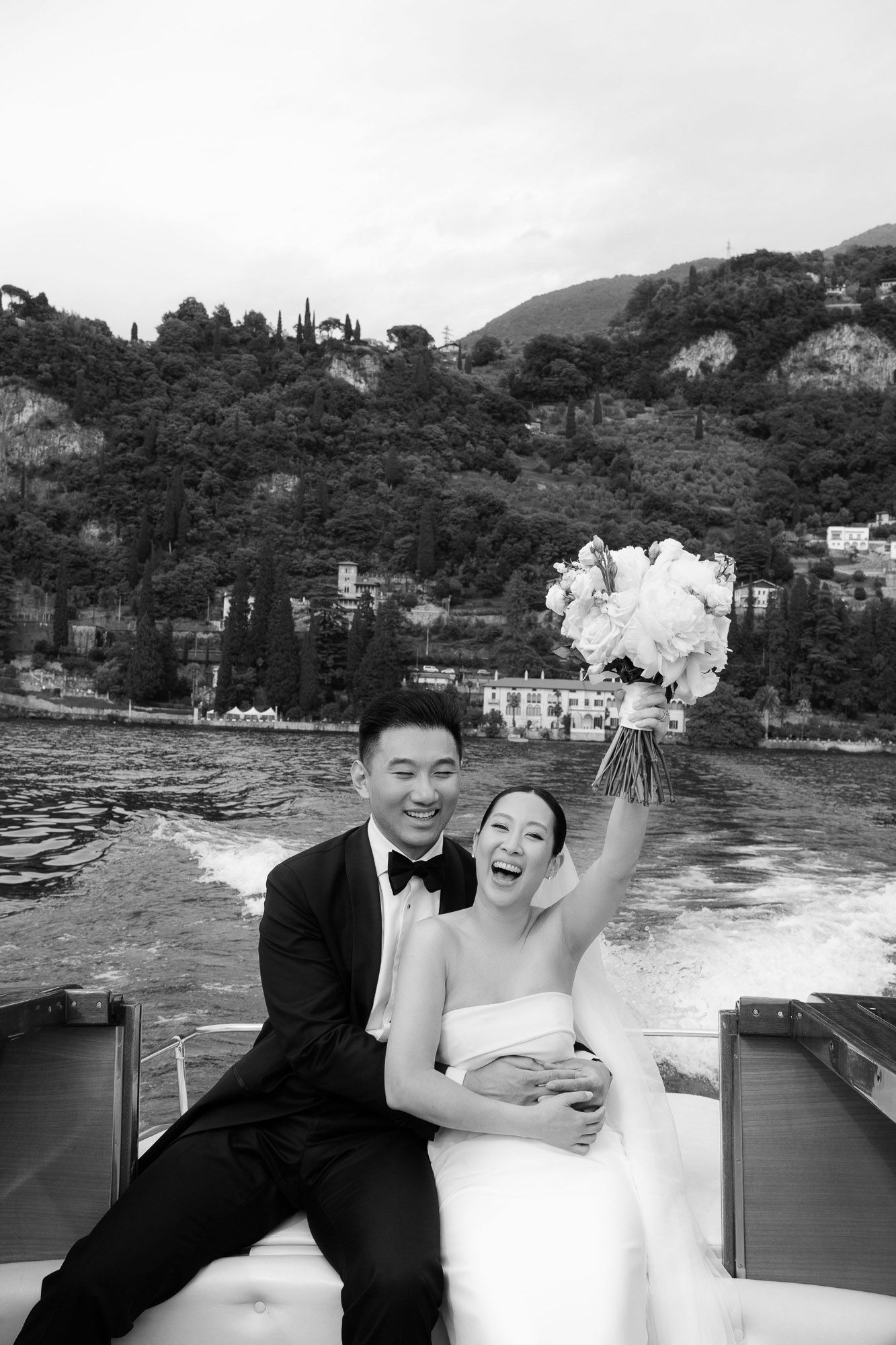 A joyful wedding couple shares a candid moment together while sitting outdoors near a scenic coastal landscape.