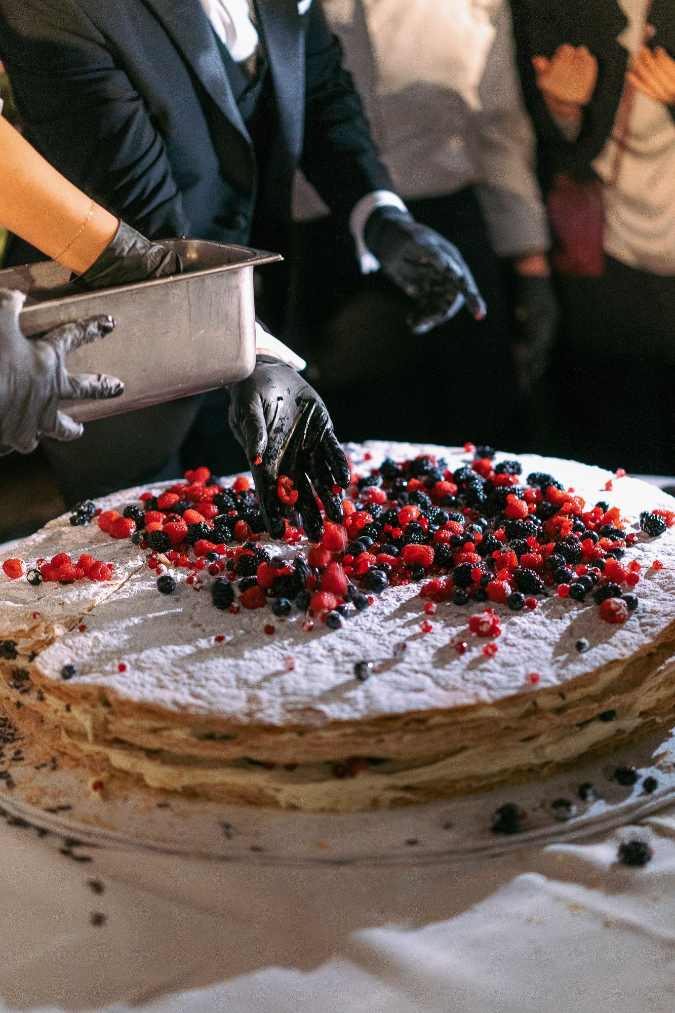 Hands slice into a large chocolate cake decorated with fresh berries and cream on a white serving platter.