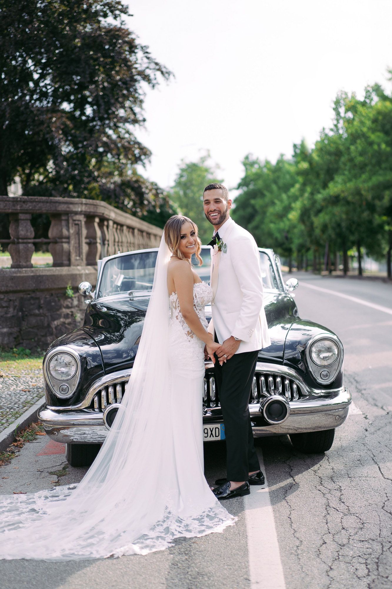 Newlywed couple poses with vintage classic car in romantic outdoor wedding photoshoot series.