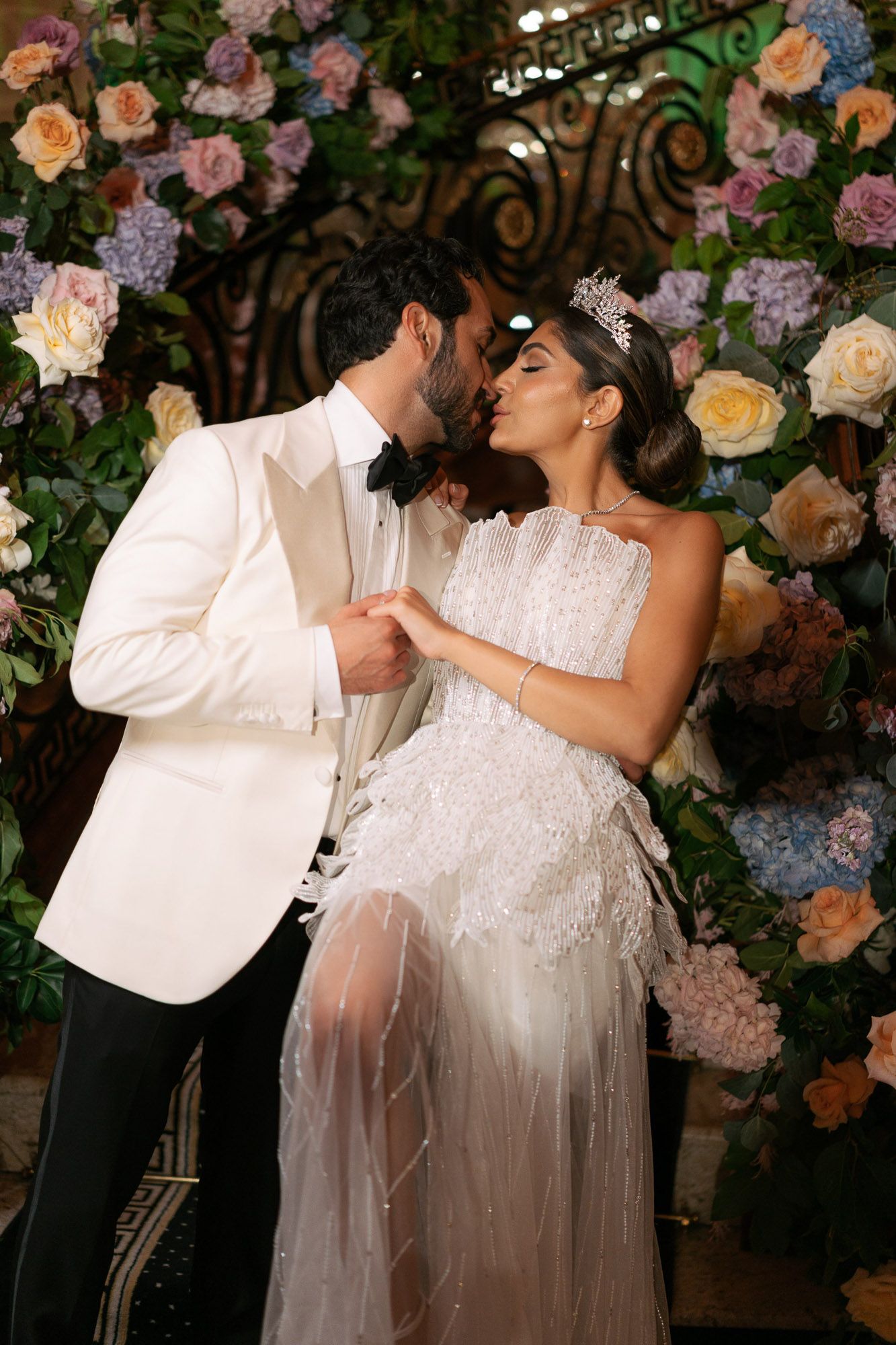 A romantic wedding moment captured under a floral arch decorated with pink and peach roses and blue hydrangeas.