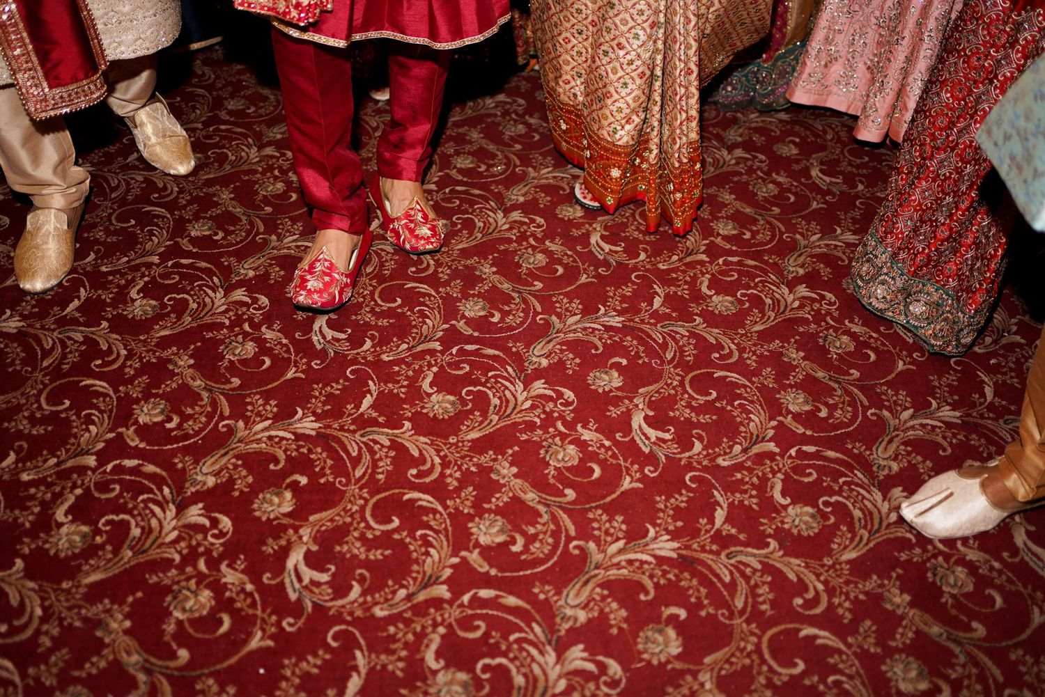 Wedding guests dance and celebrate on an ornate red patterned carpet at an elegant reception venue.