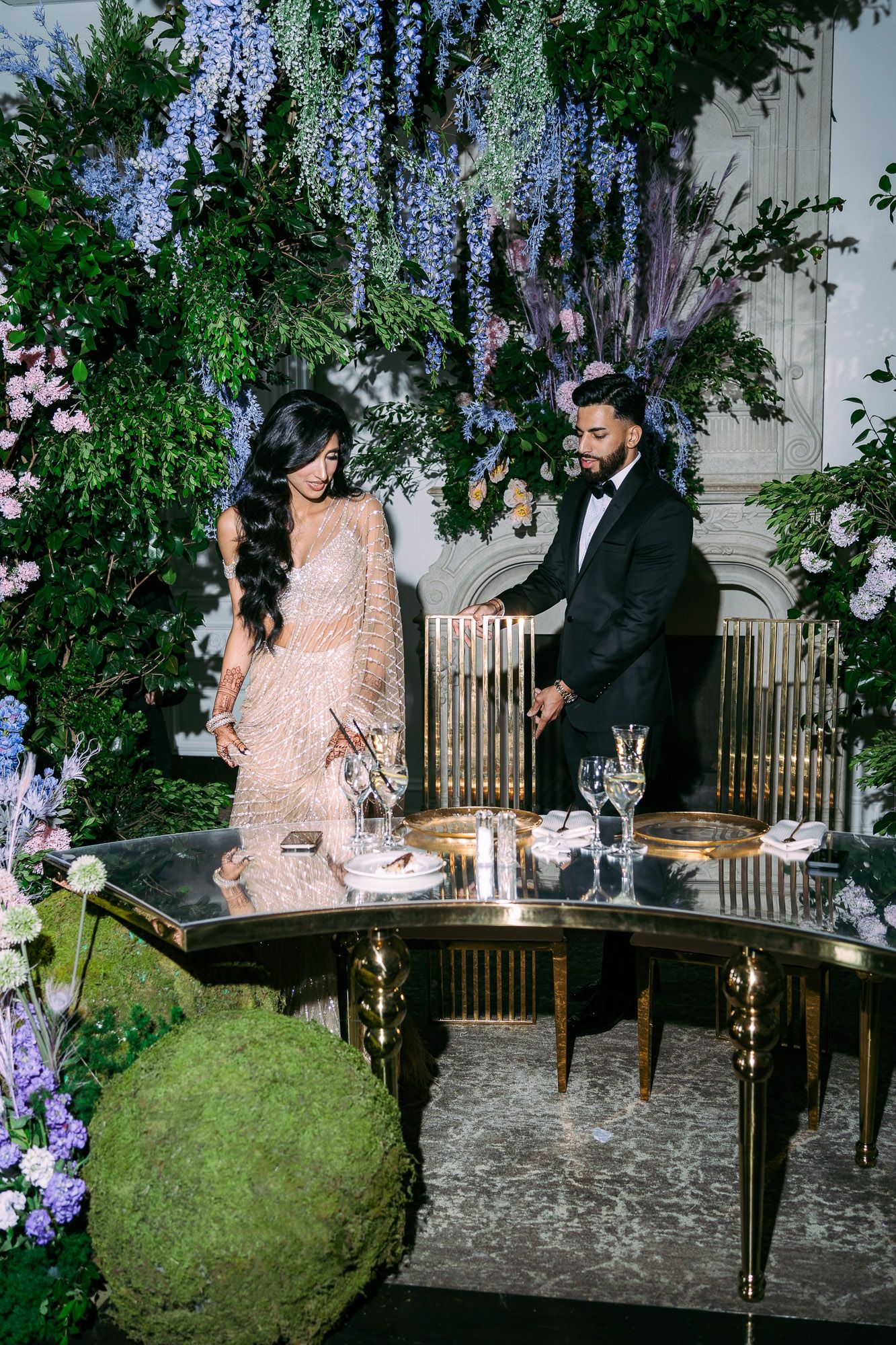 A couple sits together at an elegant outdoor venue decorated with purple wisteria and stone walls during an evening event.