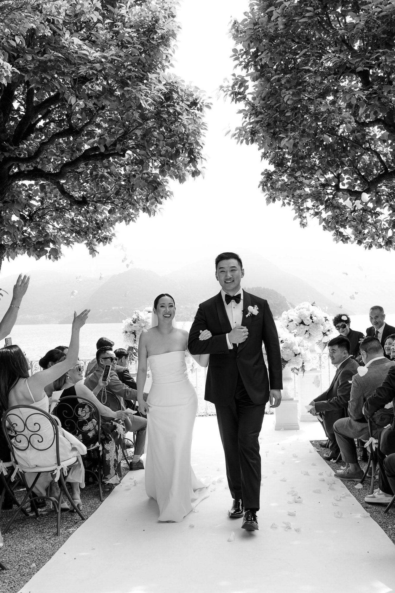A black and white panoramic sequence shows a playful moment during a wedding ceremony with guests seated outdoors.