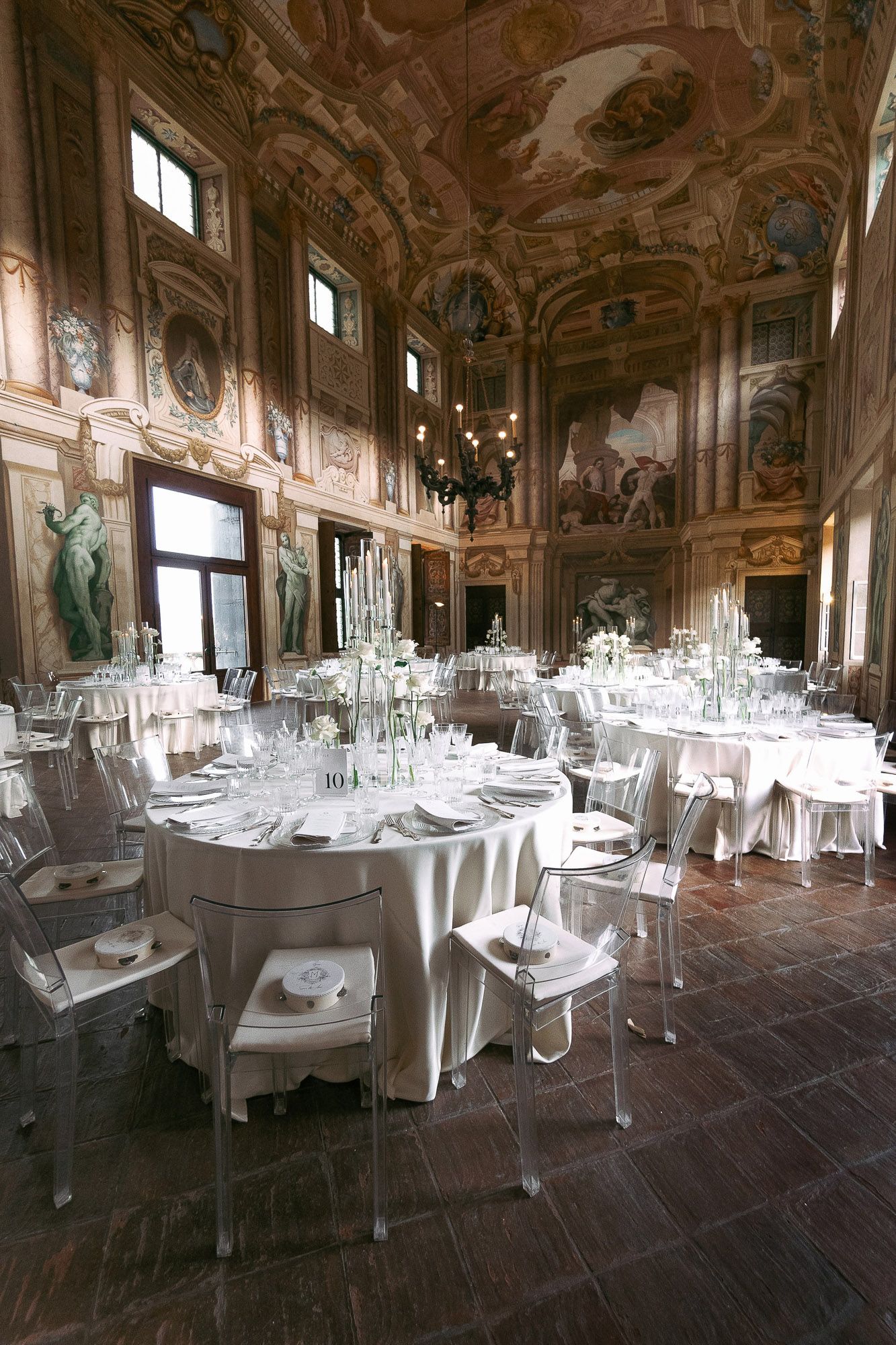Elegant banquet hall with ornate frescoed ceiling and walls set with round dining tables and ghost chairs for an event.