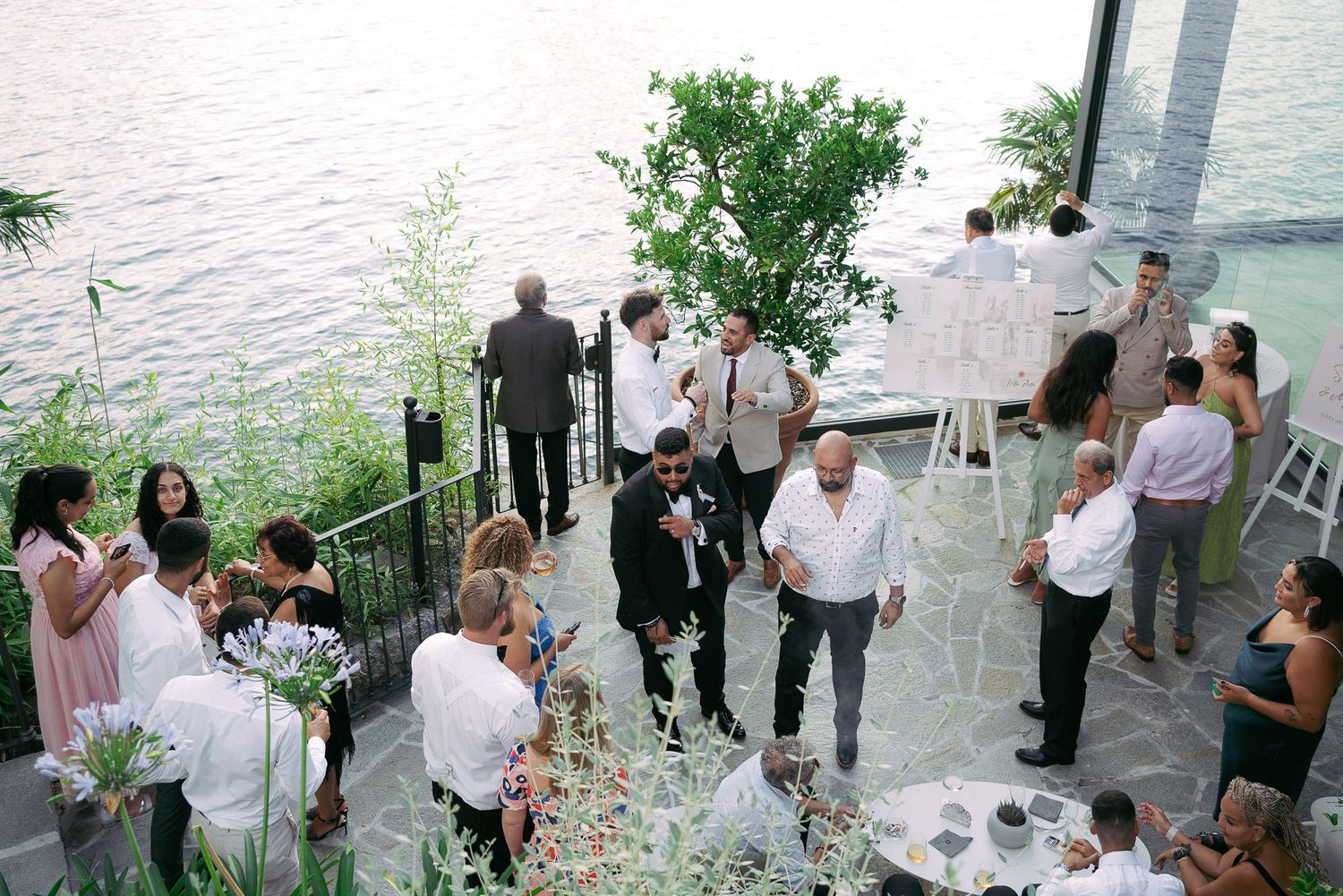 Wedding guests gather outdoors for a celebration by the waterfront with tables and chairs set up under trees.