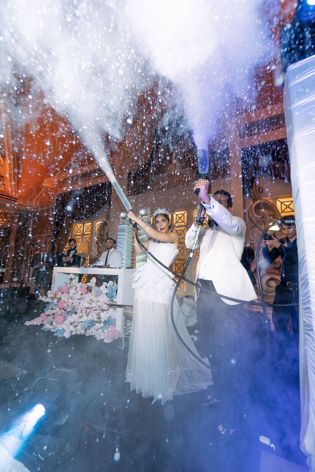 Couple spraying champagne at a wedding celebration with dramatic lighting and confetti effects in the background.