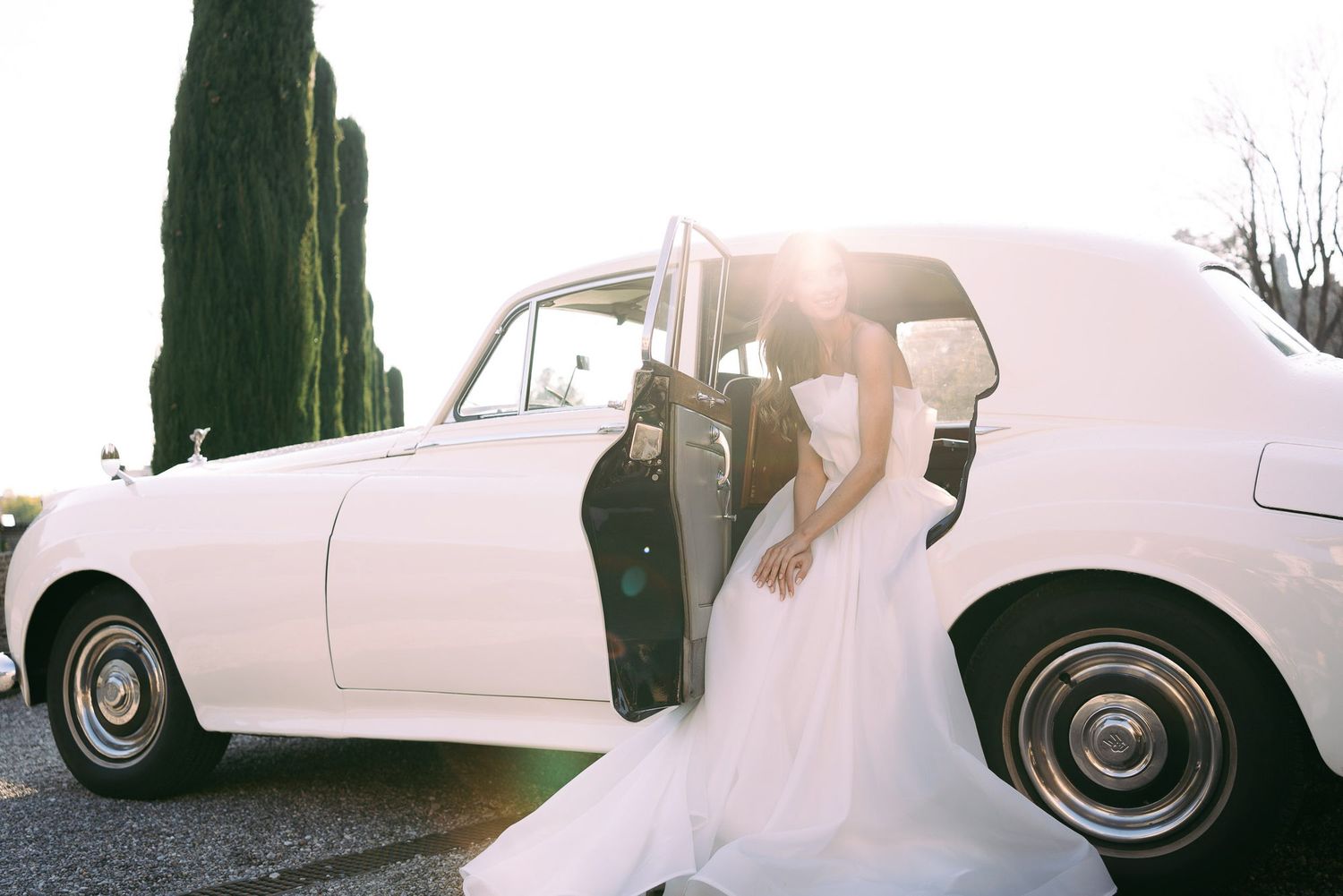 A white classic wedding car decorated with ribbons and flowers parked outside during a sunny day.