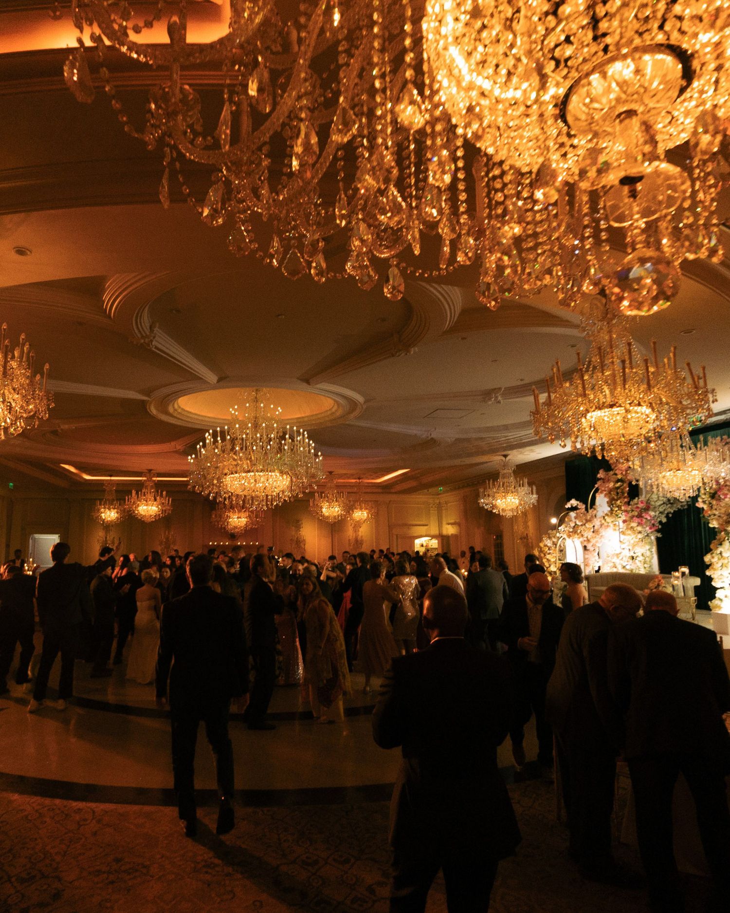 Elegant ballroom reception with glittering crystal chandeliers and large crowd of guests dancing under warm lighting.