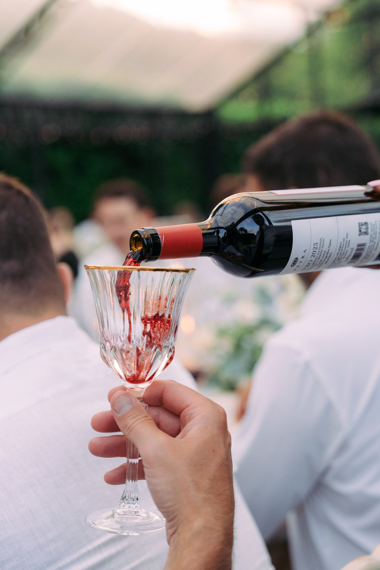 Red wine being poured into an elegant crystal glass at an outdoor gathering.