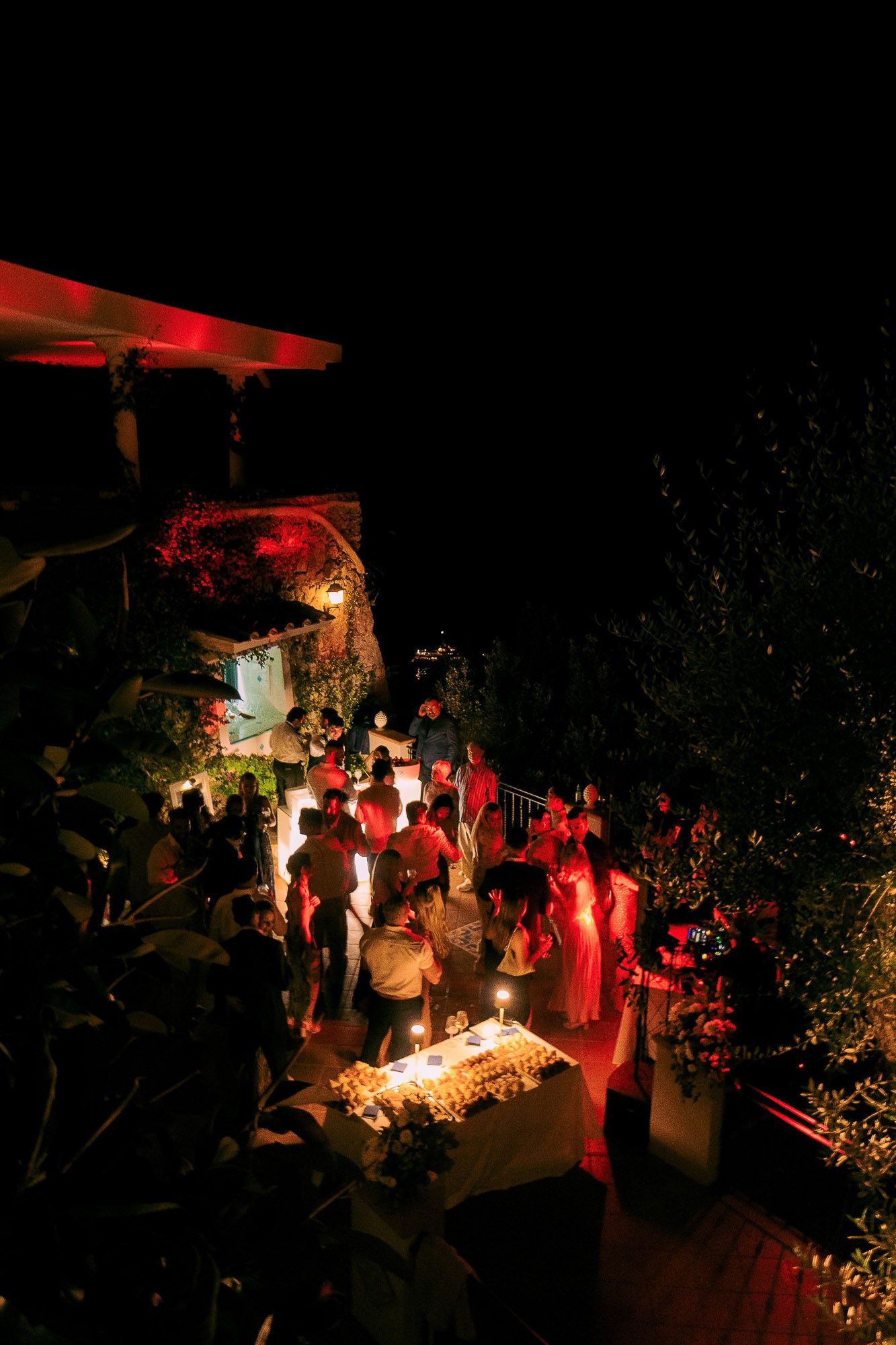 A nighttime outdoor gathering illuminated by red lights and candles creates an atmospheric party scene.