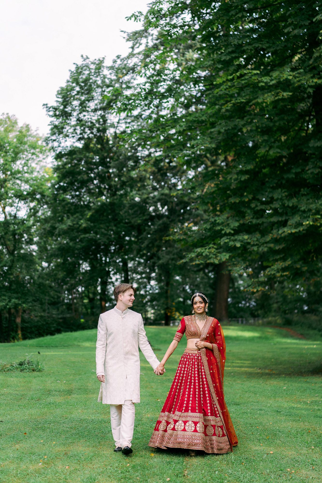 A couple in traditional wedding attire, with the bride in a red lehenga, walk together on a lush green lawn surrounded by trees.