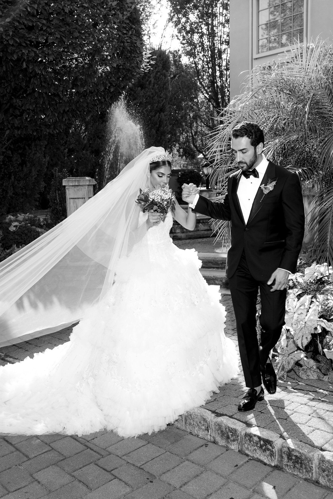 A wedding couple poses outdoors on a stone path with a flowing white gown and classic black tuxedo.