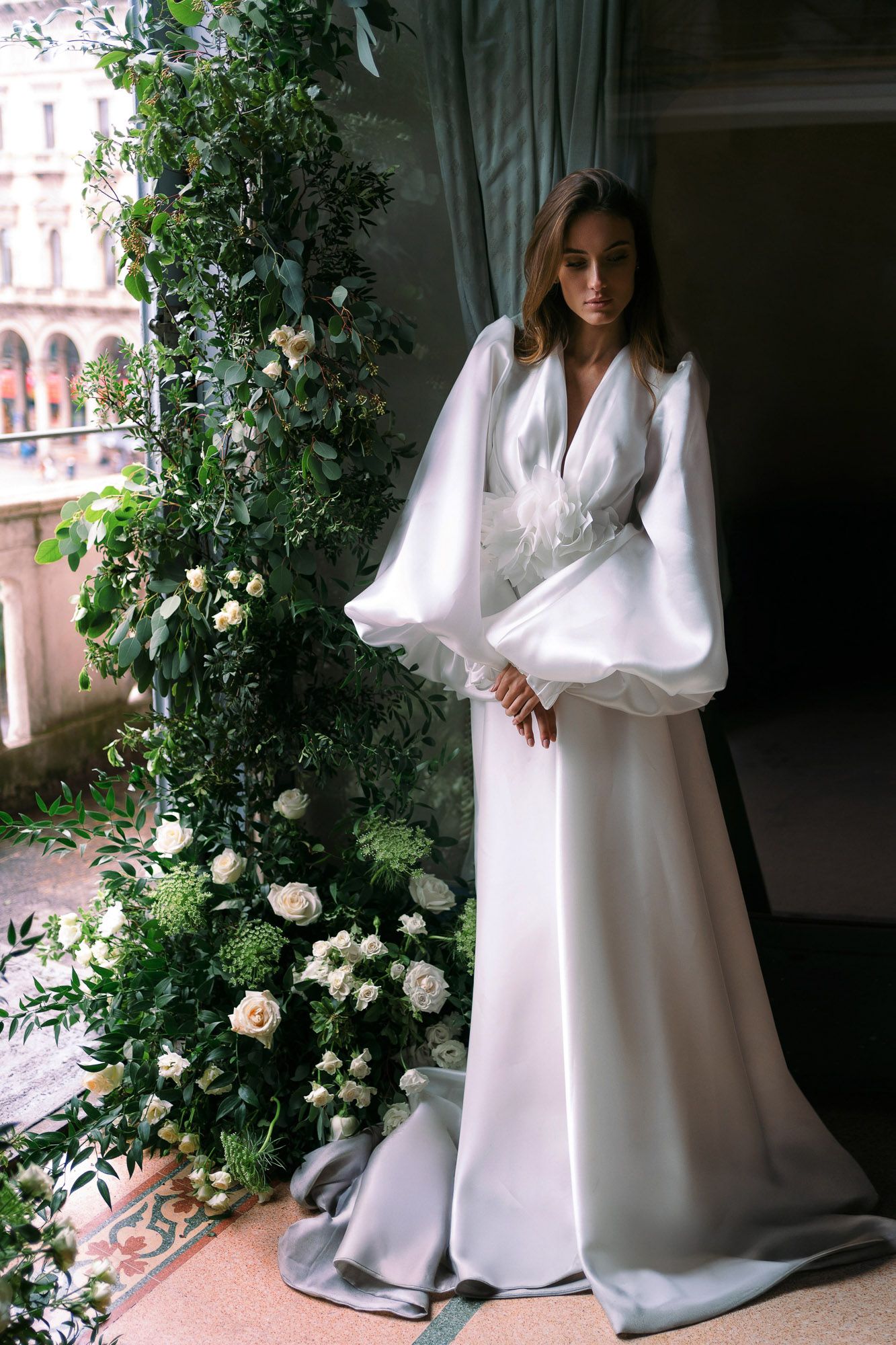 Elegant white wedding gown with dramatic balloon sleeves and flowing skirt stands beside floral archway in romantic setting.