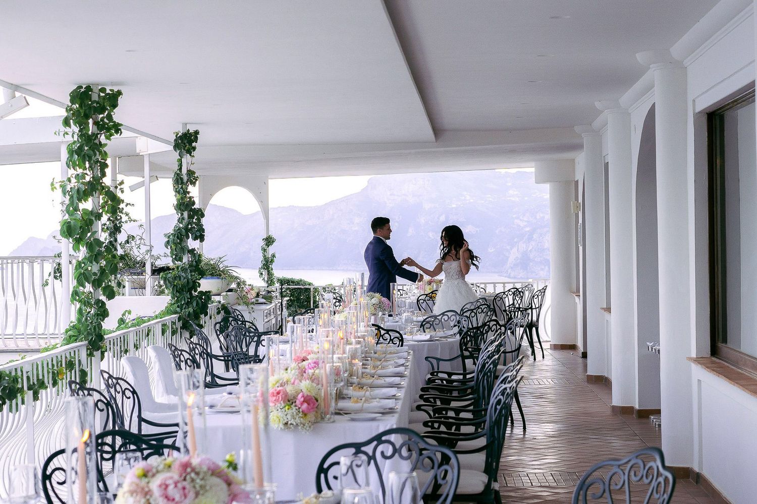 Long white covered porch with black wrought iron chairs and hanging plants creates an elegant outdoor seating area.