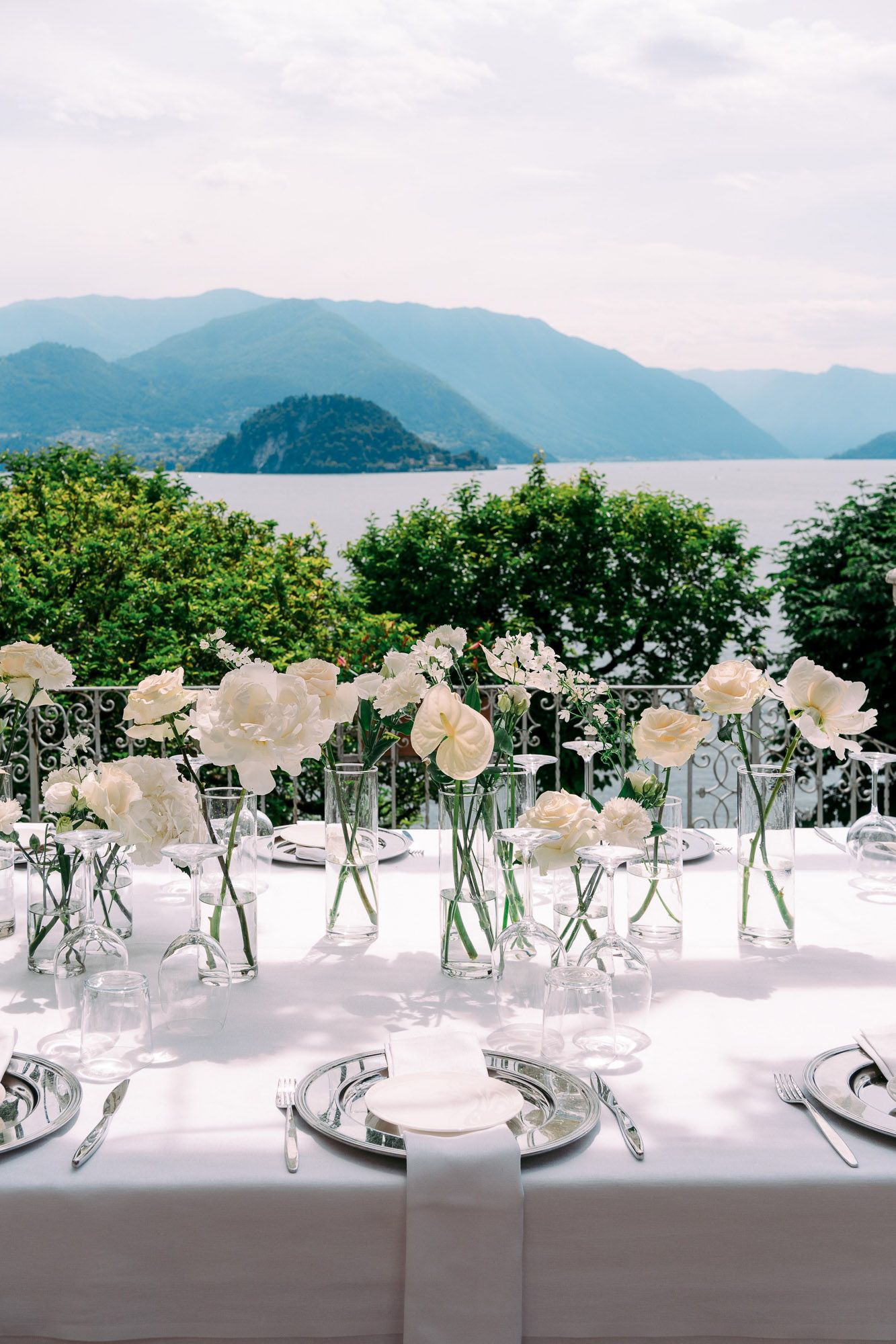 Elegant white table settings with glass vases of white flowers overlooking a scenic mountain lake view.