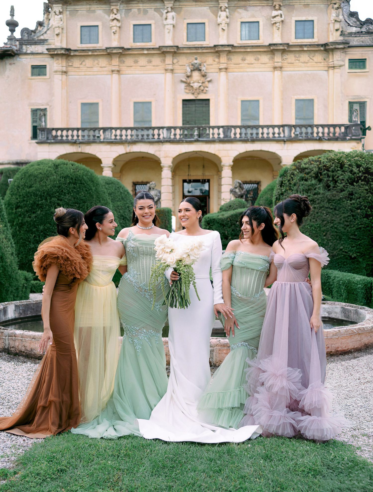 Bridesmaids in pastel dresses of mint green, yellow, and lavender pose together in a garden setting near a grand estate.