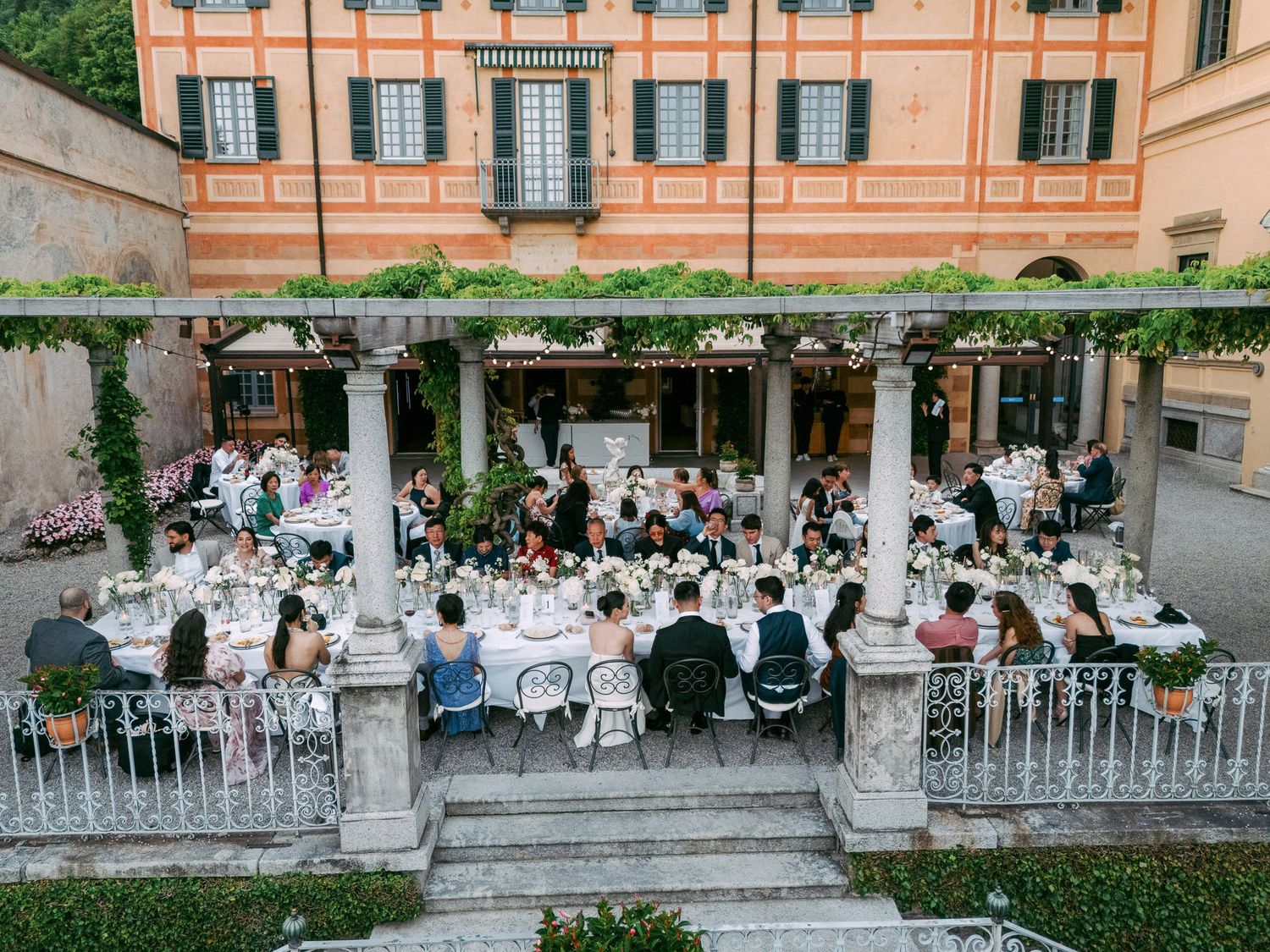 Guests seated at outdoor dining tables along a pink Mediterranean-style building with arched windows and lush greenery.