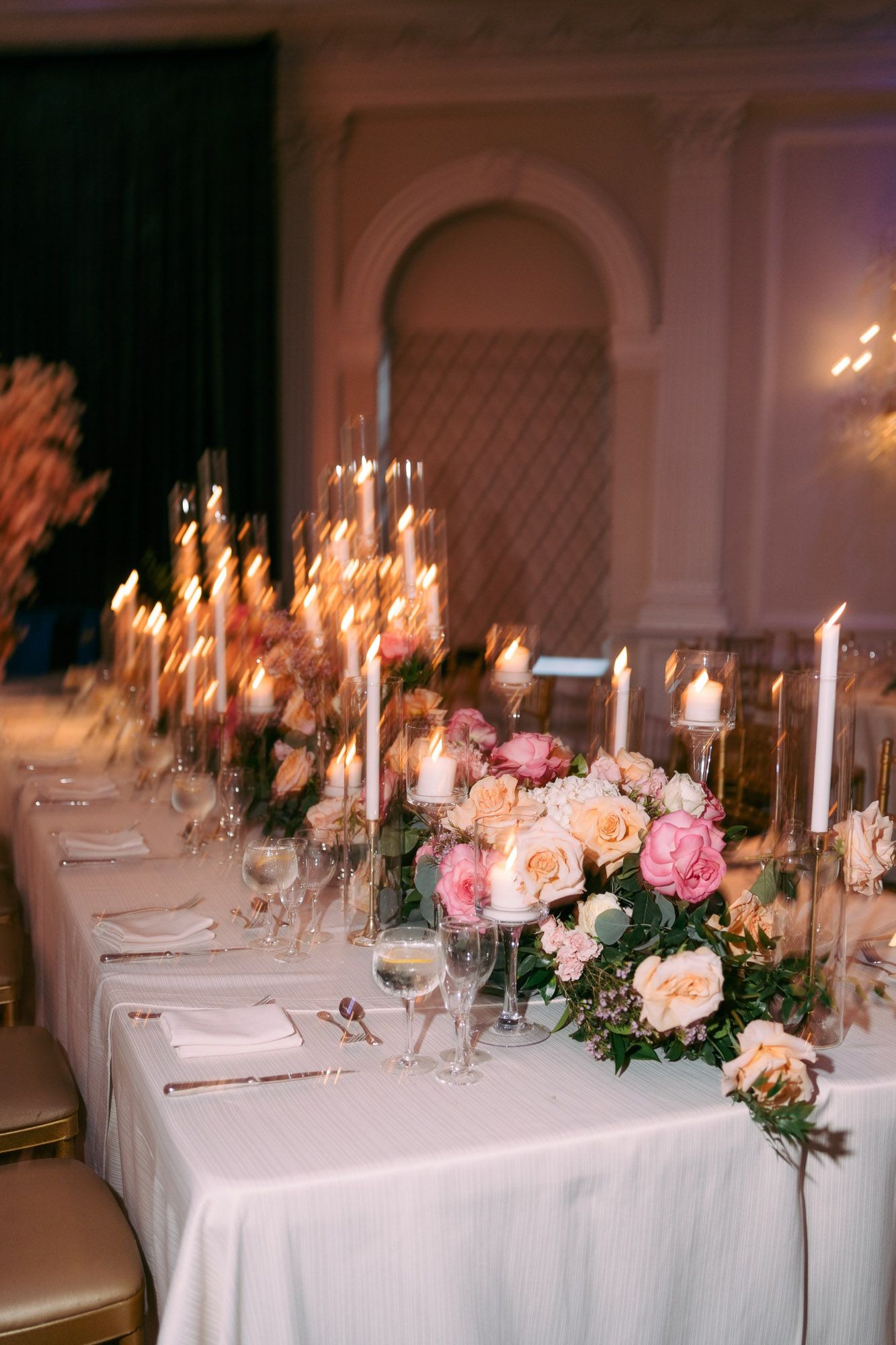 Elegant wedding reception tables decorated with white linens, pink roses, and glowing taper candles in a romantic indoor setting.