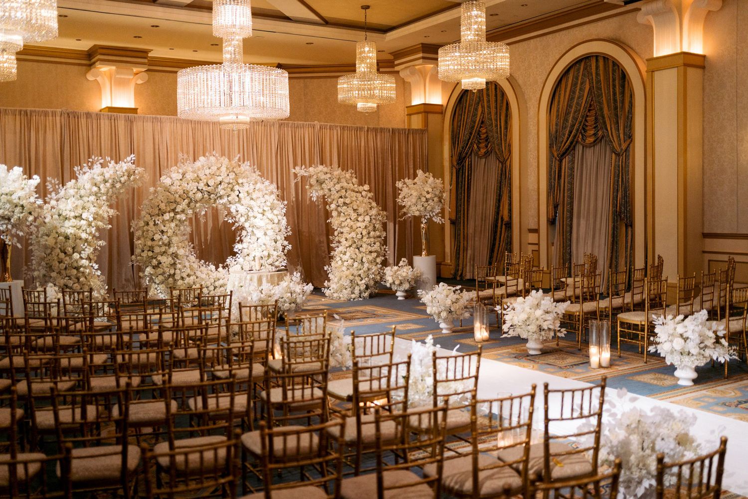 Elegant wedding ceremony aisle decorated with white cherry blossom trees and crystal chandeliers in a luxurious ballroom.