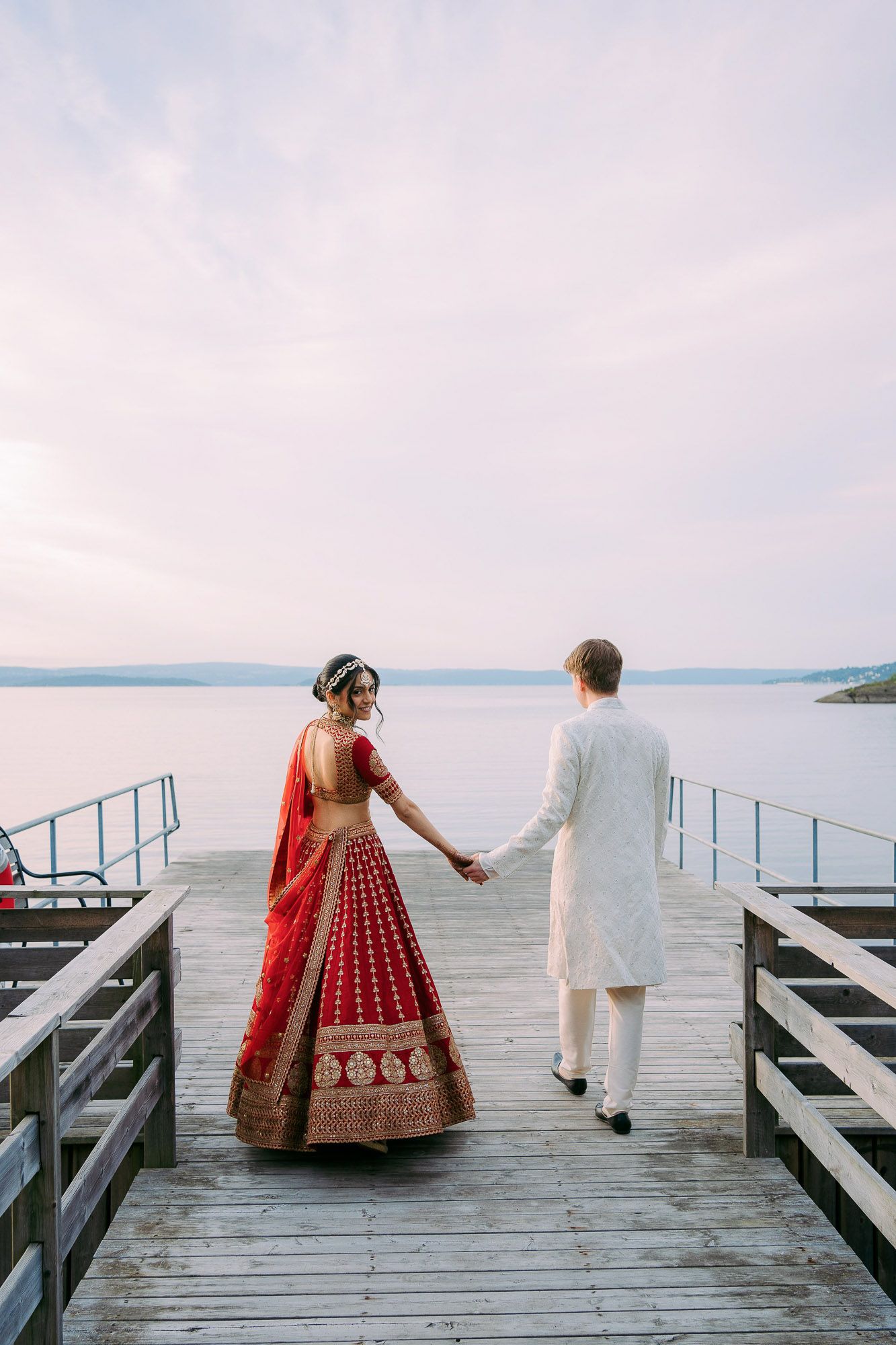 A couple stands on a wooden pier overlooking a serene lake during sunset in a romantic wedding photoshoot.