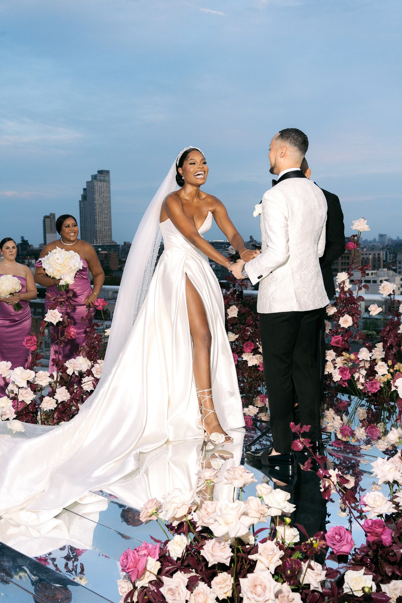 A wedding couple poses on a rooftop with pink and white floral decorations against a city skyline backdrop.