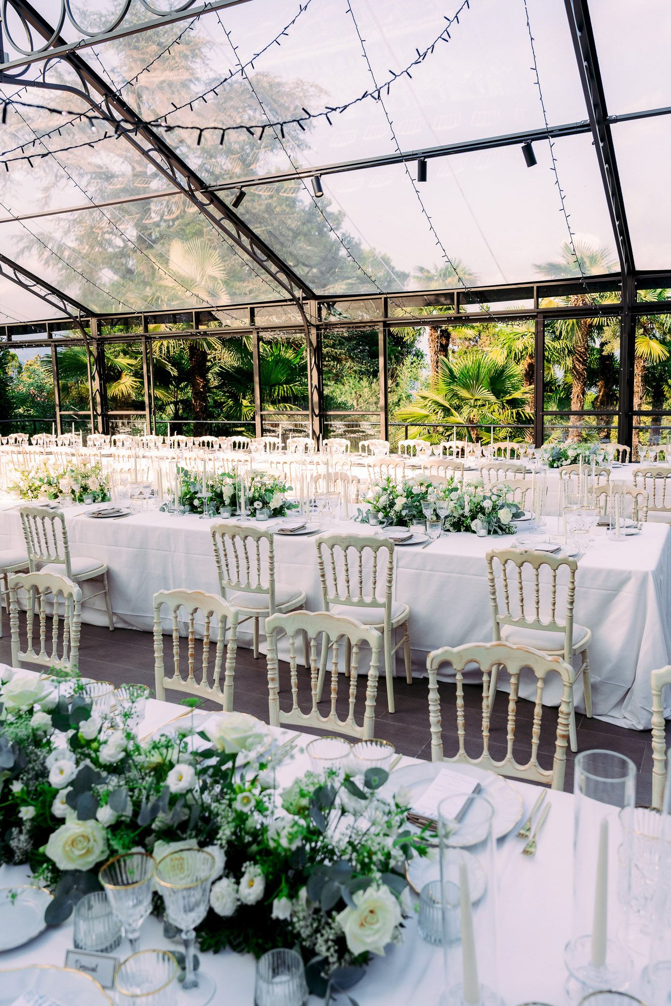 Elegant outdoor wedding reception setup with white tables, chiavari chairs and floral arrangements under a clear tent canopy.