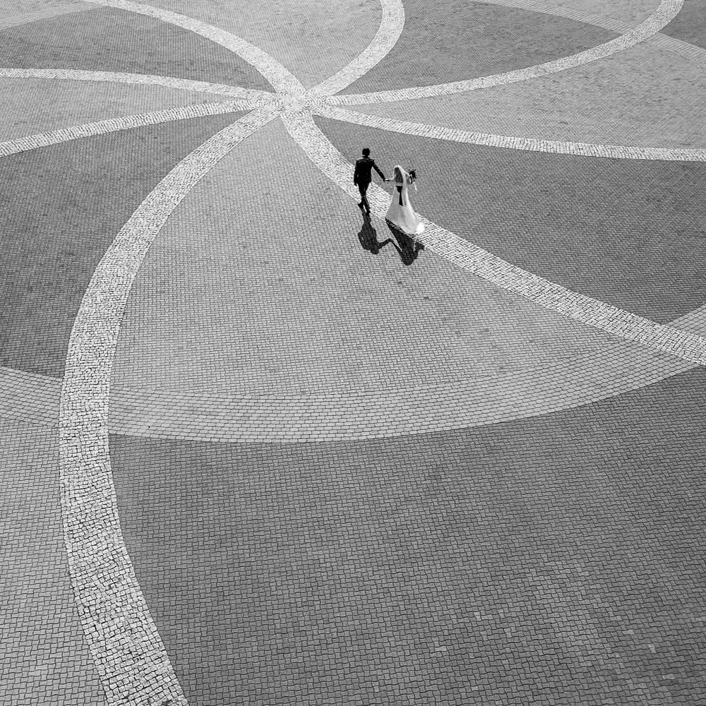 Two figures cast long shadows while walking across a geometric patterned plaza with curved white lines on grey pavement.