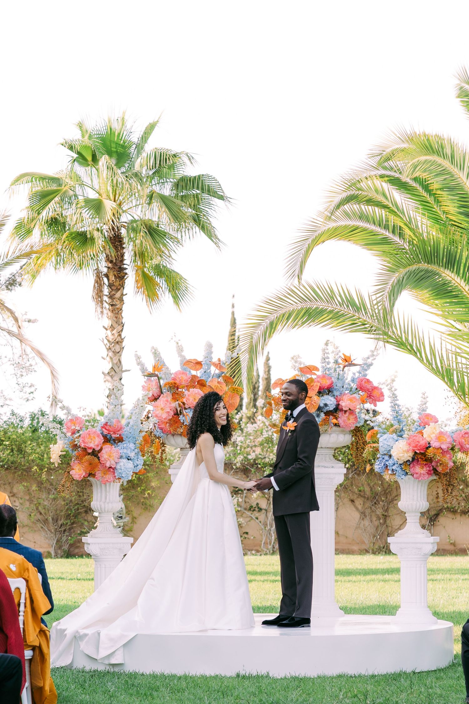 Elegant outdoor wedding ceremony setup with floral arrangements and white decor under palm trees.