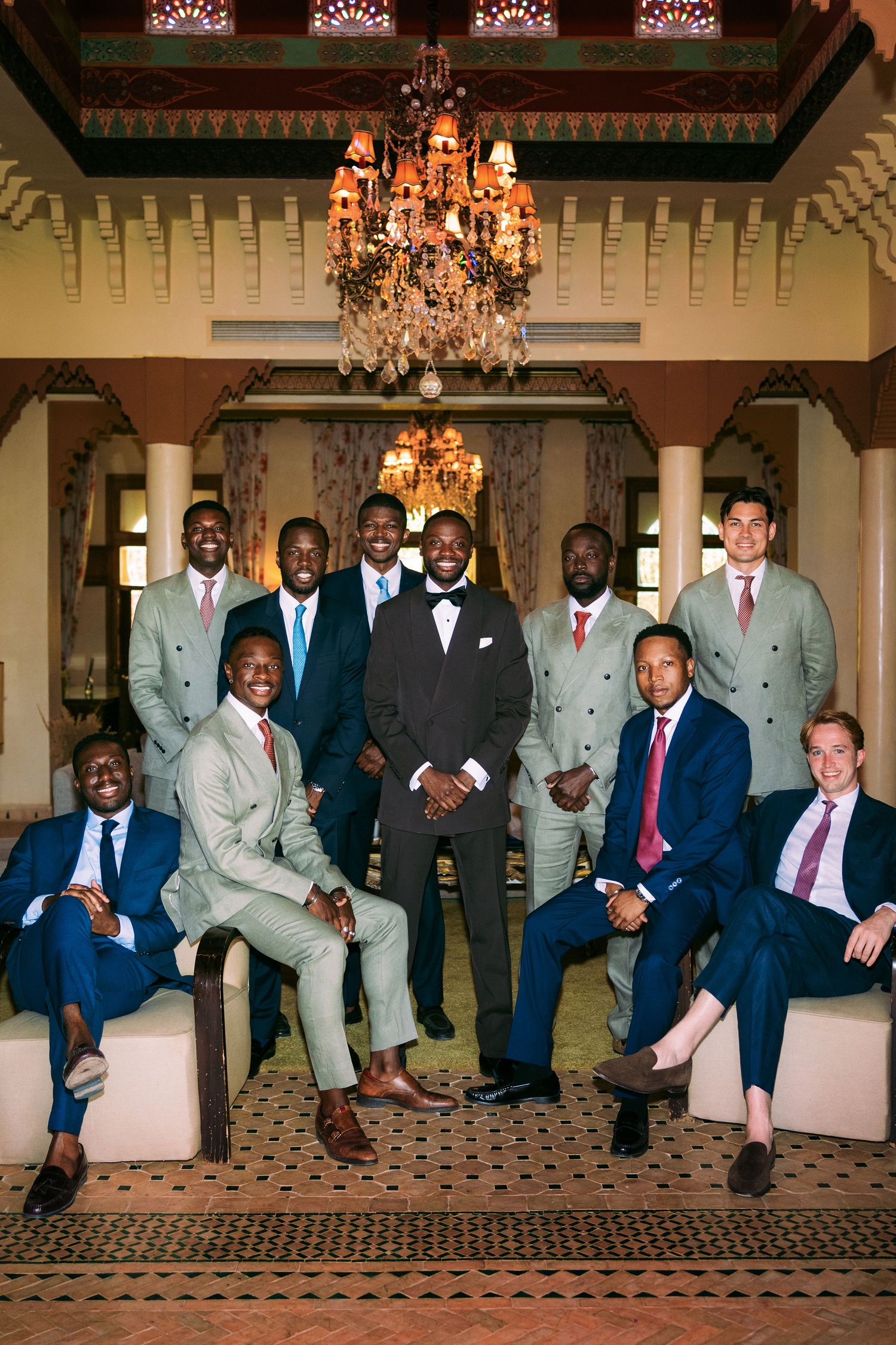 Wedding party groomsmen in suits pose together on elegant couch in formal room with chandeliers.