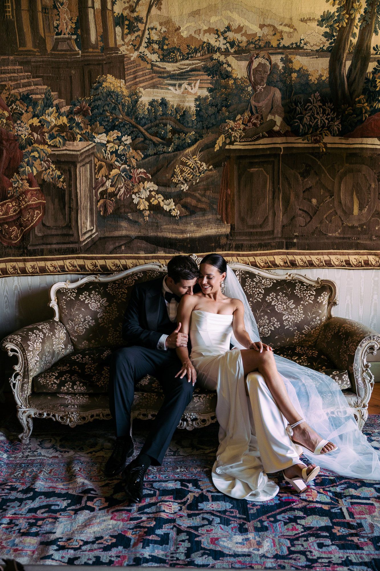 Romantic couple lounges together on an ornate vintage sofa with rose petals scattered on an antique rug.