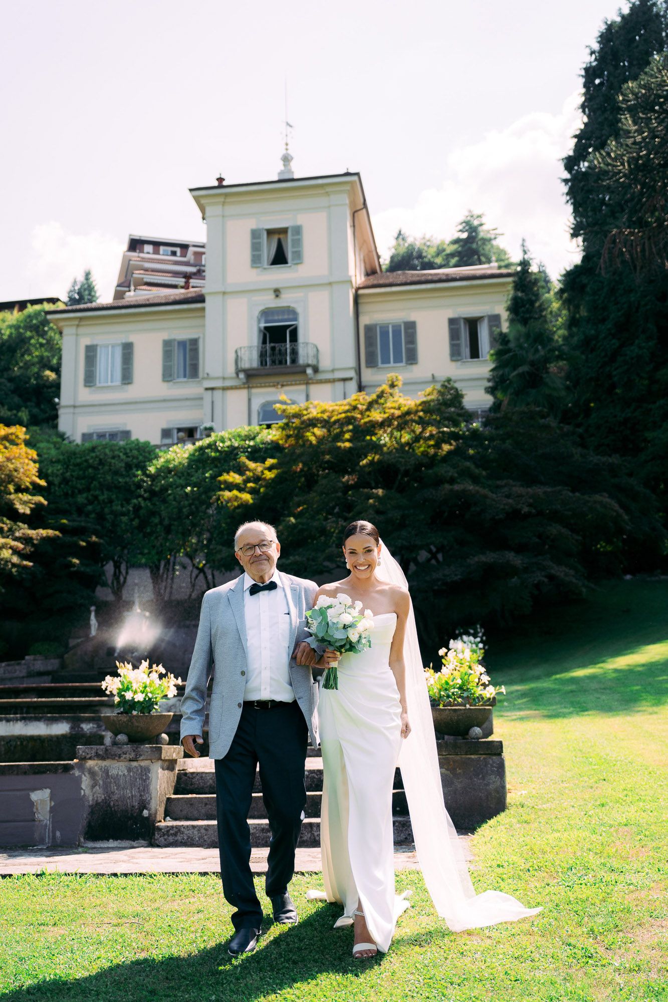 Elegant wedding couple walks together on a sunny day across a manicured lawn near a white mansion.