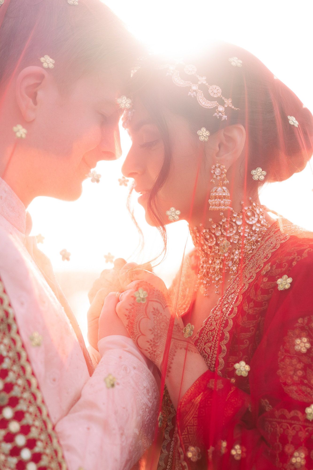 Silhouetted figures in traditional red Chinese wedding attire stand together in warm sunlit haze with delicate floral details.