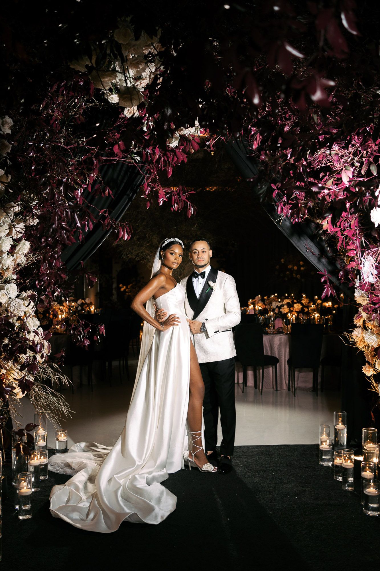 Elegant wedding couple poses under dramatic floral arch with pink blossoms and moody lighting at evening reception.