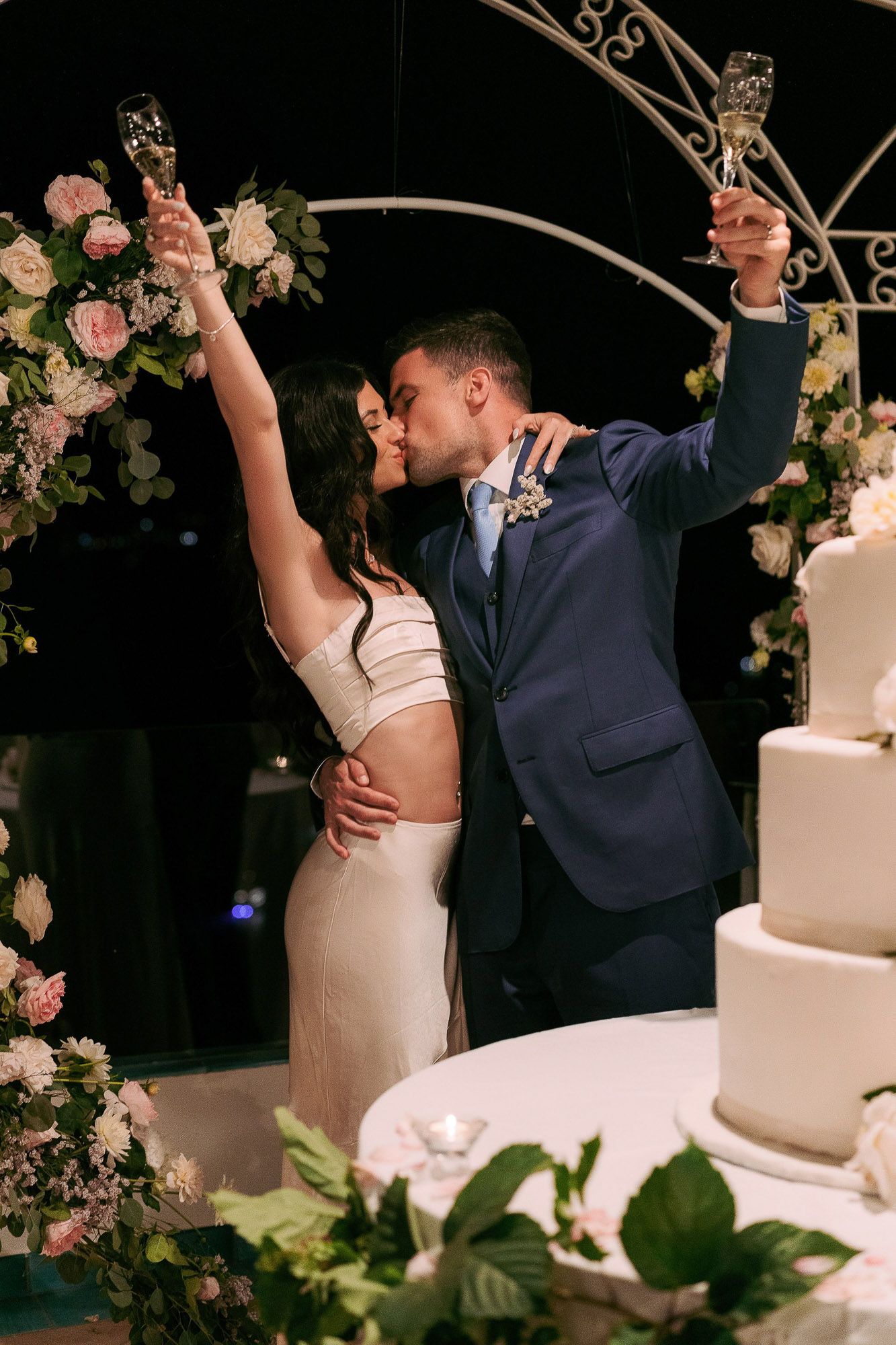 A joyful wedding couple celebrates by the cake, surrounded by floral decorations and an elegant arch.
