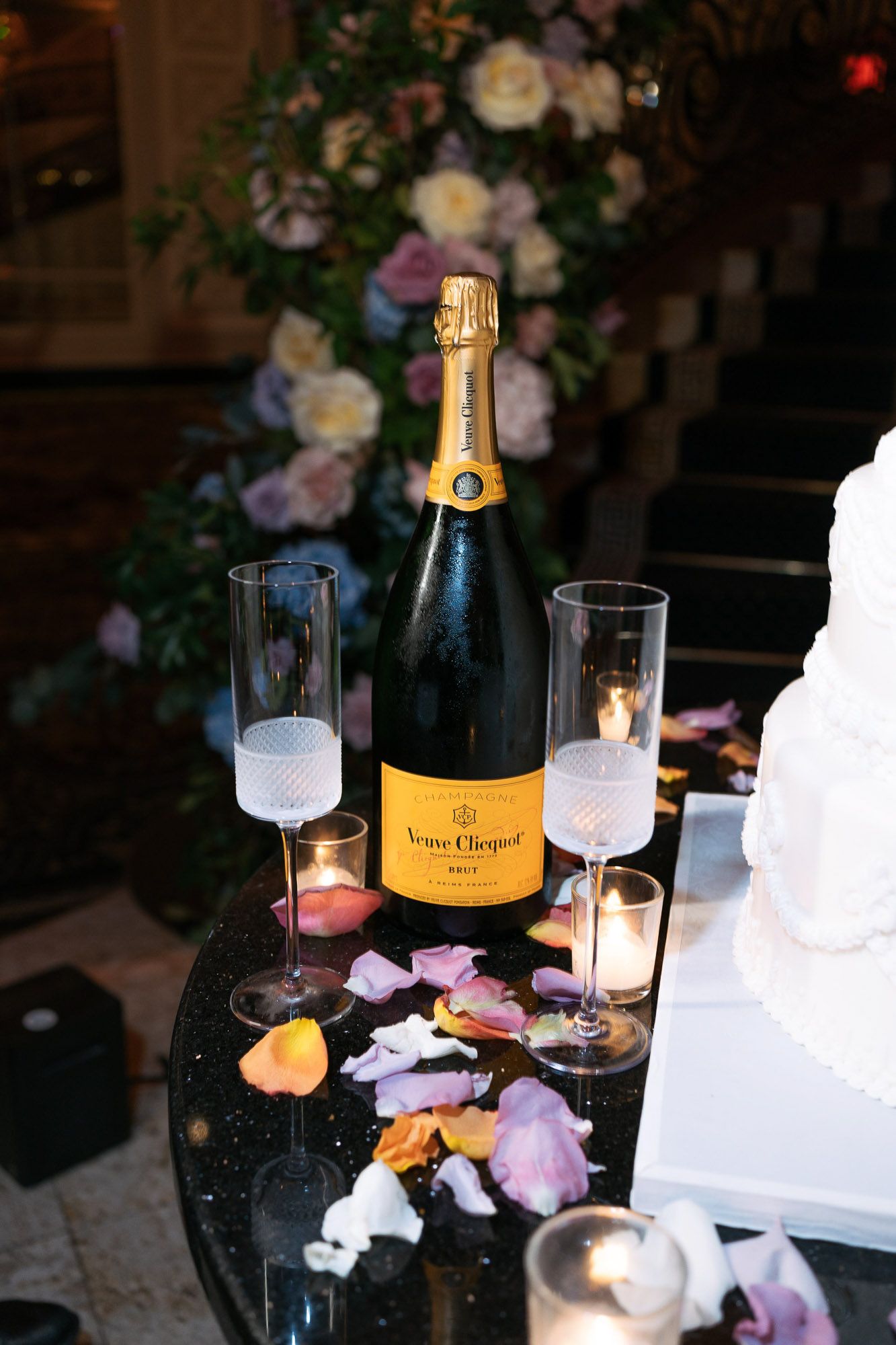 Veuve Clicquot champagne bottle and glasses displayed on a dark table with scattered rose petals and candlelight.