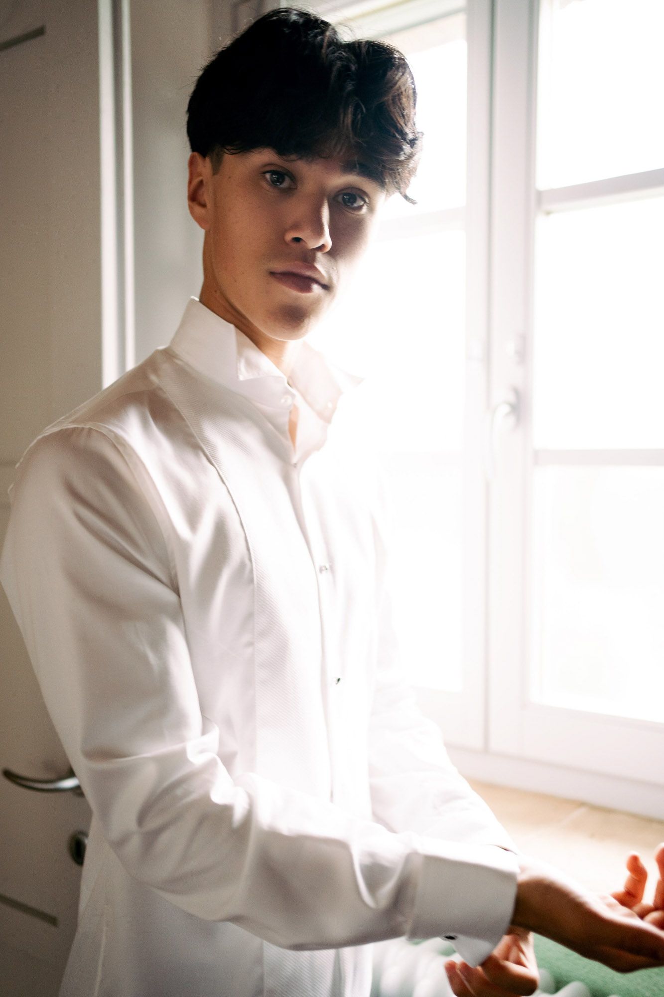 Portrait of a person in a crisp white button-down shirt standing by a sunlit window.