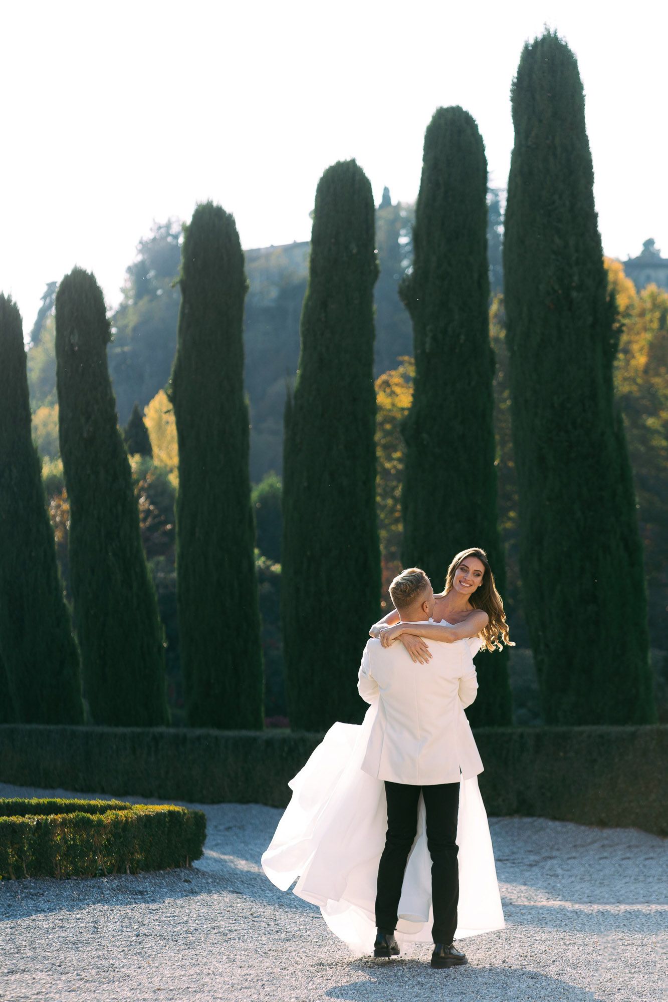 Romantic wedding couple dancing and twirling along a cypress tree-lined path in a formal garden setting.